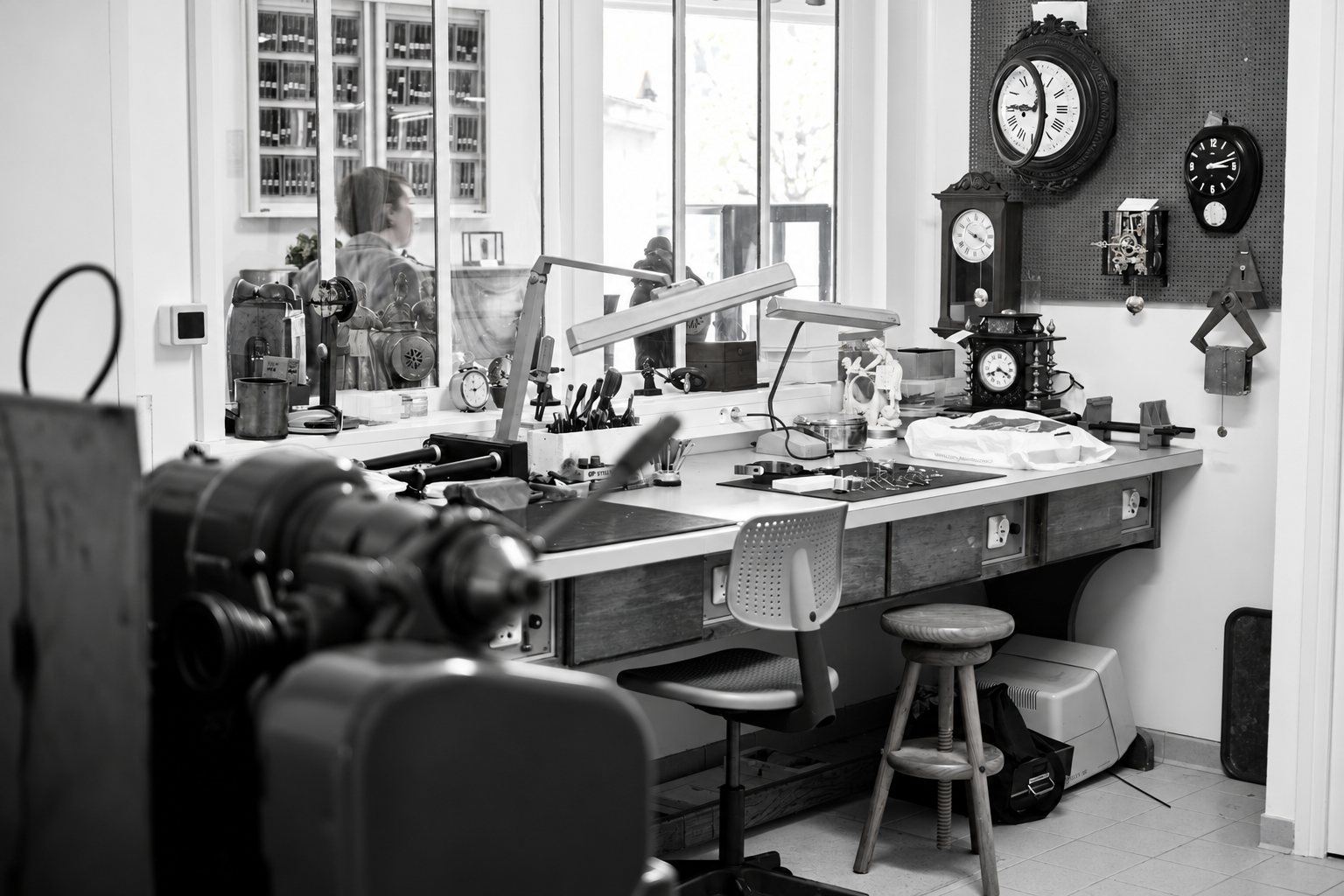 Photo en noir et blanc d'un atelier d'horlogerie avec un établi équipé de lampes et d'outils, un tabouret, une chaise et plusieurs pendules anciennes accrochées au mur.