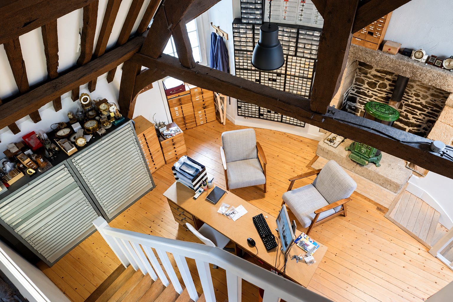 Vue plongeante d'un atelier d'horloger sous charpente apparente, avec bureau en bois, deux fauteuils, meubles à tiroirs et pendules anciennes exposées.