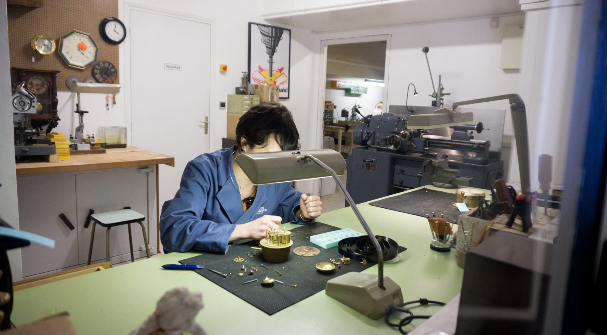 Un horloger en blouse bleue travaille sur un mécanisme de pendule sous une lampe, à un établi vert, dans un atelier équipé d'un tour et d'horloges murales.