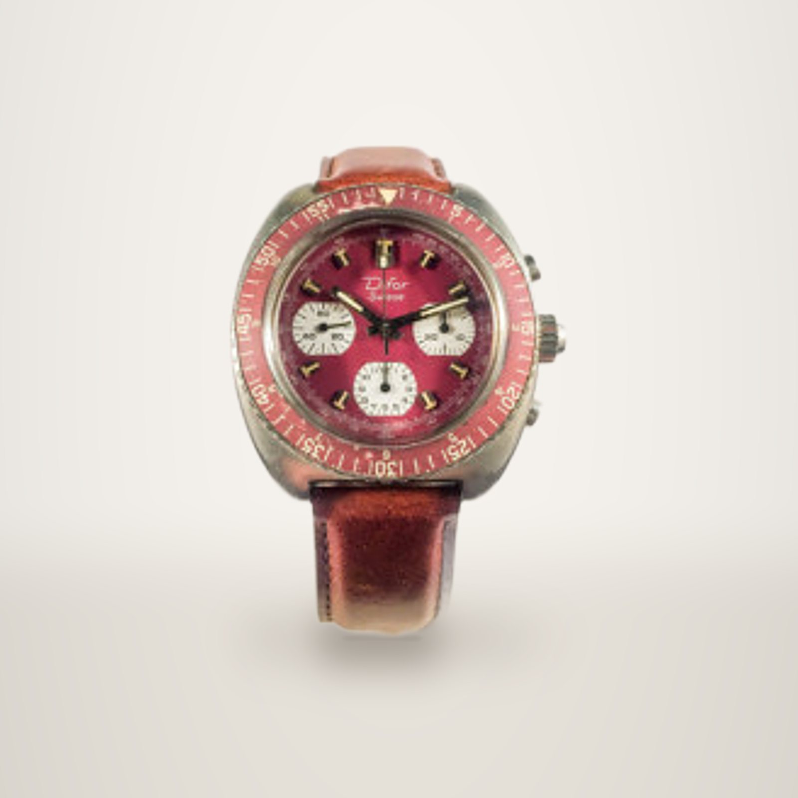 Montre chronographe ancienne avec cadran rouge, trois compteurs blancs et bracelet en cuir marron, posée sur un socle blanc devant un fond uni.