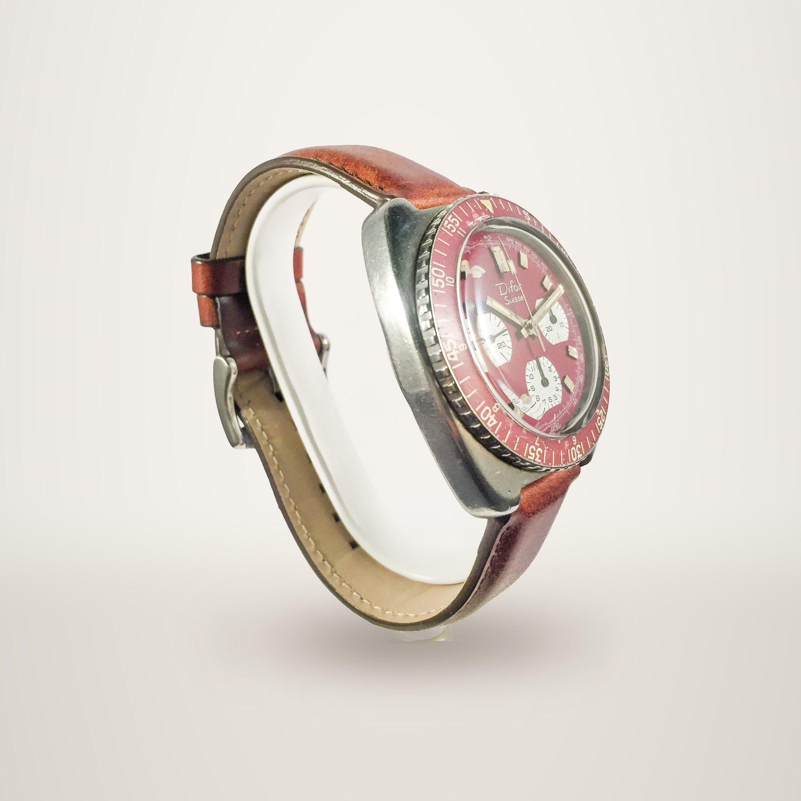 Montre chronographe ancienne de marque Difor avec cadran et lunette rouges, boîtier en acier et bracelet en cuir brun, posée sur un support blanc.