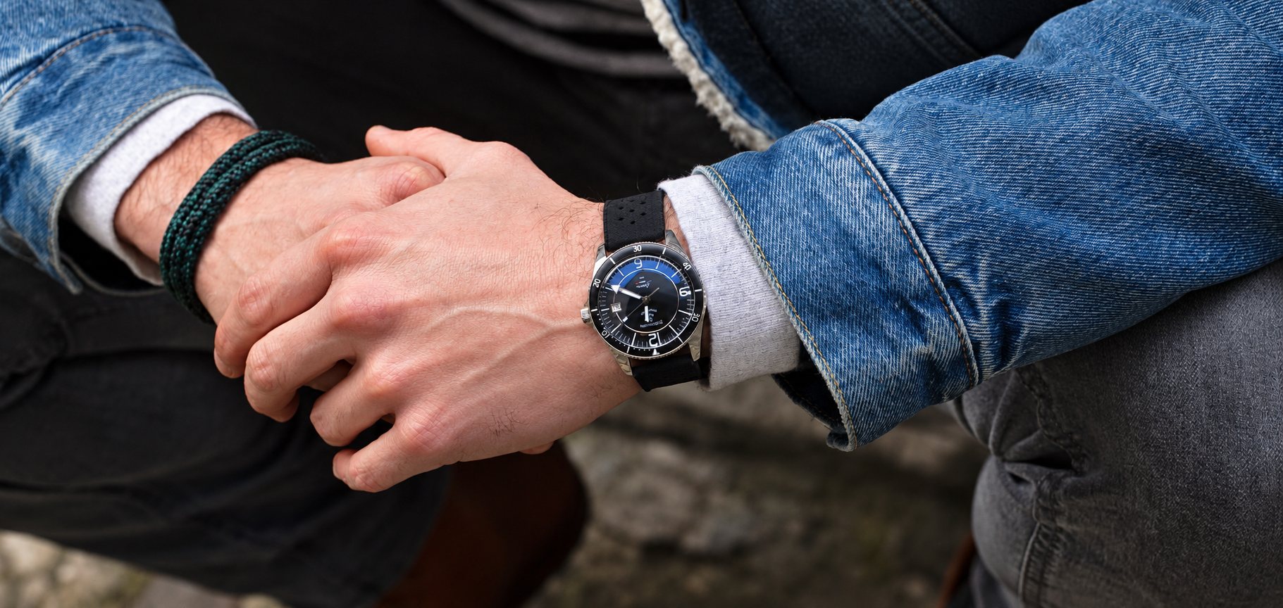 Gros plan sur les mains jointes d'un homme en veste en jean, portant une montre de plongée à cadran bleu et bracelet noir.