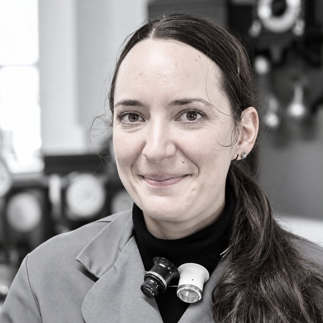 Portrait en noir et blanc d'une femme souriante en blouse, portant deux loupes d'horloger autour du cou, devant des horloges floues.